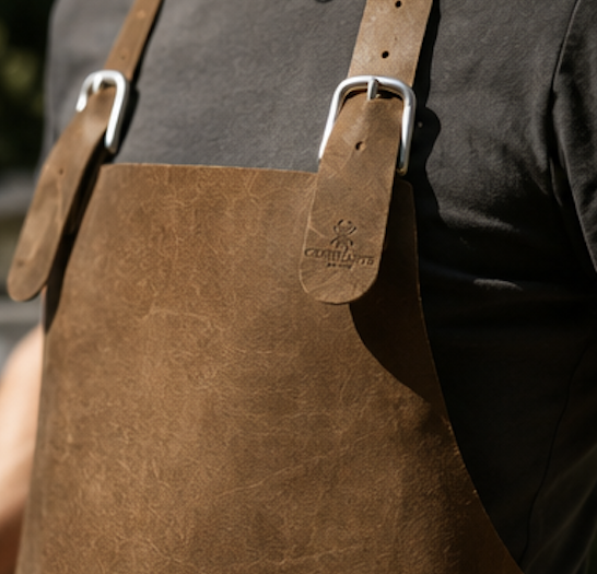 Adult wearing a brown leather apron with metal buckle straps over a dark tee, midsection visible.