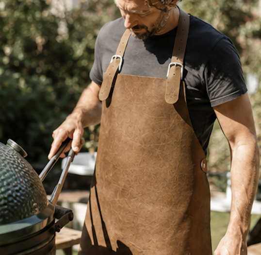 Man wearing a brown leather apron grilling outdoors, tending a kettle-style grill in a sunny backyard.